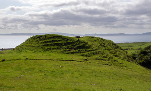 A grass-covered burial cairn with dents that look like steps leading up to its top on a field close to the sea