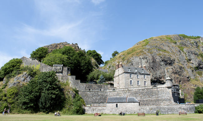A grassy park in front of the high stone walls of Dumbarton Castle, with the Governor's House visible beyond and the prominent Dumbarton rock dominating the landscape.