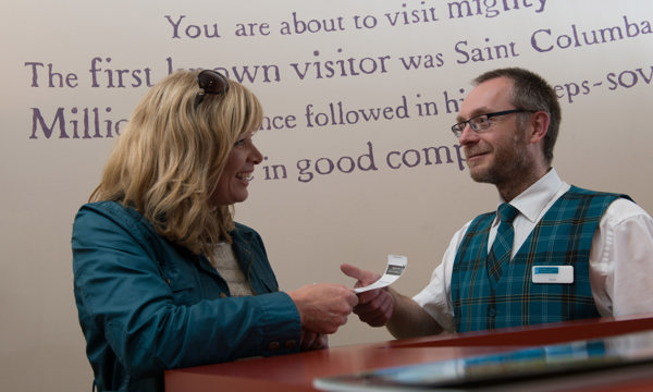 Male member of staff in Historic Scotland tartan uniform accepting a tickets from a lady in a green jacket and sunglasses on her head