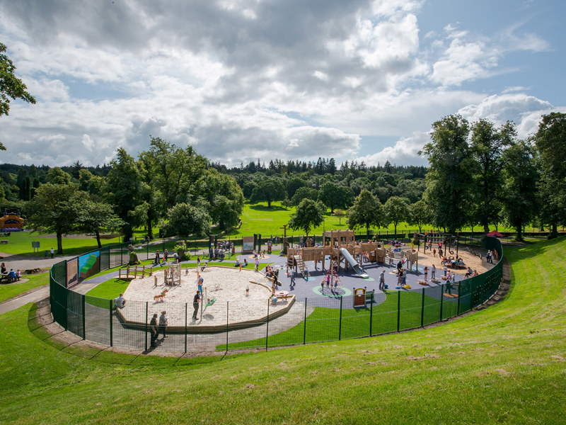 A playground full of children on a sunny day. The playground is surround by a fence and appears to be situated in the middle of a much large park filled with trees.