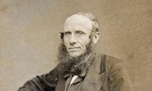 black and white portrait photograph of a person seated with their right arm resting on a table and left hand holding a piece of paper. They are wearing a suit and overcoat with a large bow tie, and they have a long, wispy beard.