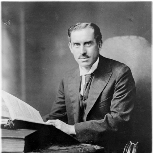 Black and white photograph of a person wearing a suit and tie seated at a table. They are looking into the camera and holding a large book in their hands, as if reading.