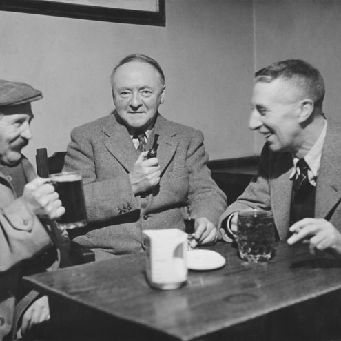 Black and white photo of three people seated at a table inside a public house. All are drinking and smiling, as if sharing a joke, and the person in the centre is looking at the camera and holding a pipe.