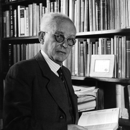 Black and white portrait photograph of a person standing in a library holding an open book. They are wearing a dark suit and tie and round glasses, and are looking directly at the camera.