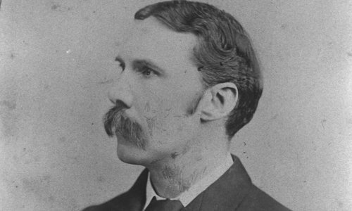 Black and white portrait photo of a person looking to the left, wearing a dark suit and tie. They have a large handlebar moustache.
