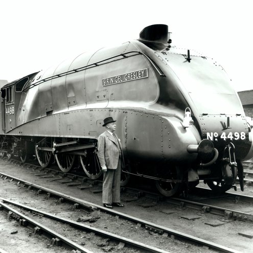 Black and white photograph of a person in a grey suit and dark trilby hat standing next to, and looking up at, a steam locomotive engine in a goods yard.