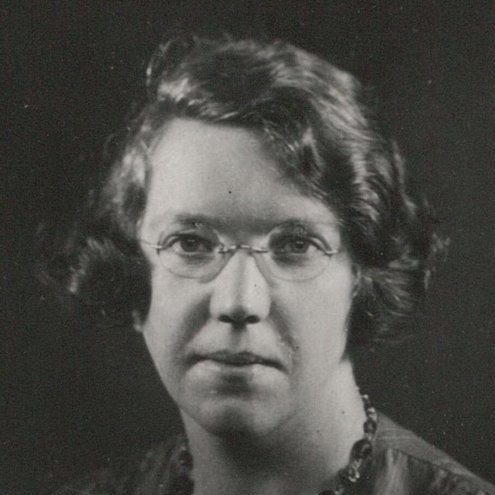 Black and white headshot portrait of a person wearing wire-rimmed glasses and a beaded necklace over a dark top.