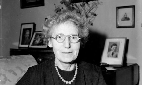 Black and white portrait photograph of a person sitting with their arms folded in an armchair. They are wearing glasses and a necklace, and behind them on a sideboard table are family photographs and a bunch of flowers in a vase.