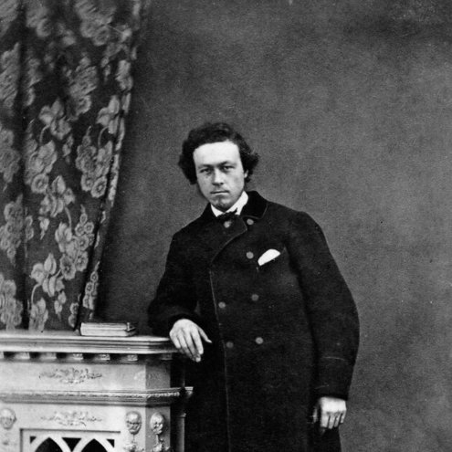 Black and white image of a man wearing formal attire of the mid 19th century, leaning on a lectern which has a closed book on it. 