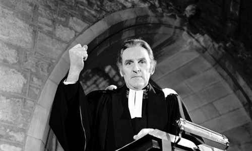 Black and white portrait photo of a person dressed as a priest and pretending to give a sermon. They are standing in a pulpit, looking down at the camera with their right arm raised into a fist as if strongly talking about something.