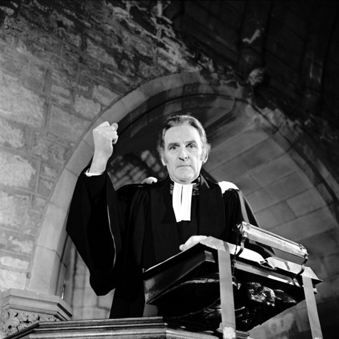 Black and white portrait photo of a person dressed as a priest and pretending to give a sermon. They are standing in a pulpit, looking down at the camera with their right arm raised into a fist as if strongly talking about something.