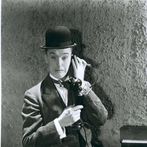 Black and white portrait photo of a person wearing a suit and bowler hat, pretending to talk on an old fashioned telephone. They have a sad or confused looking expression on their face.