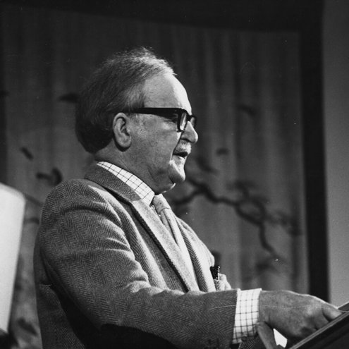 Black and white photo of a person giving a reading, standing at a lectern. They are wearing a jacket and tie and thick-rimmed black glasses. There is a book open in front of them.