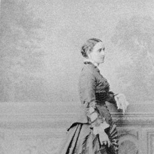 Black and white photograph of a person standing in profile, facing to the right. They are wearing a large dark dress of expensive-looking fabric and are holding a fan in their right hand.