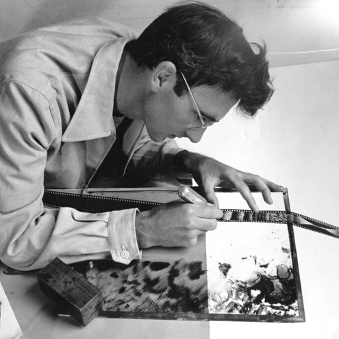 Black and white photo of a person in a shirt and glasses leaning over a table. They are marking a series of film negatives with a pen, as light shines upwards from underneath.