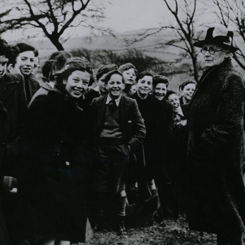 Black and white photo of a group of 1950s schoolchildren and a person supervising them. They are at a tree planting ceremony, and the uniformed children are laughing and smiling. The person is looking sternly at them, with their hands in their coat pockets.