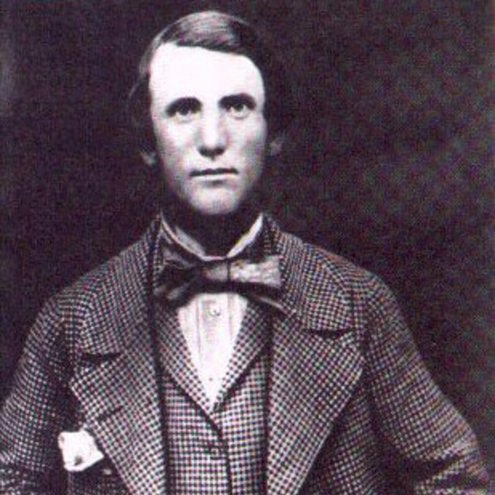Black and white portrait photo of a person wearing a tweed suit and waistcoat. They also have a large dark bow tie and are looking at the camera with a relaxed expression, their right hand holding the lapel of their jacket.