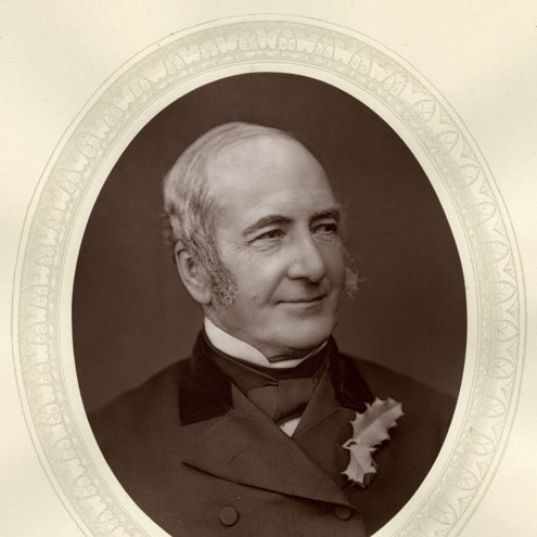 An early black and white portrait photo of a person looking off to the right. They are smiling, and wearing a dark suit, white shirt and black cravat, with two holly leaves pinned to their lapel. They have large grey sideburns reaching down to their cheeks.