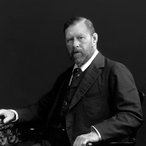 Black and white image of a seated man wearing a suit and tie. He has one hand on the arm of the chair and is looking at the camera.