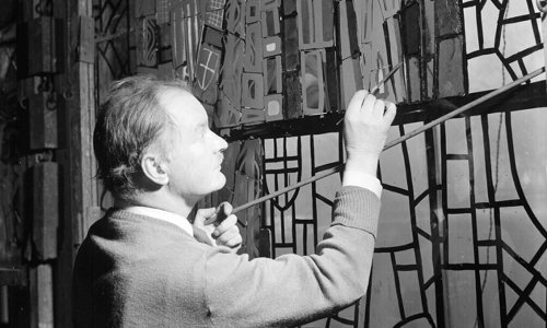 Black and white photograph of a person working on a very large stained glass window. They are carefully painting a section of the glass with a fine brush.