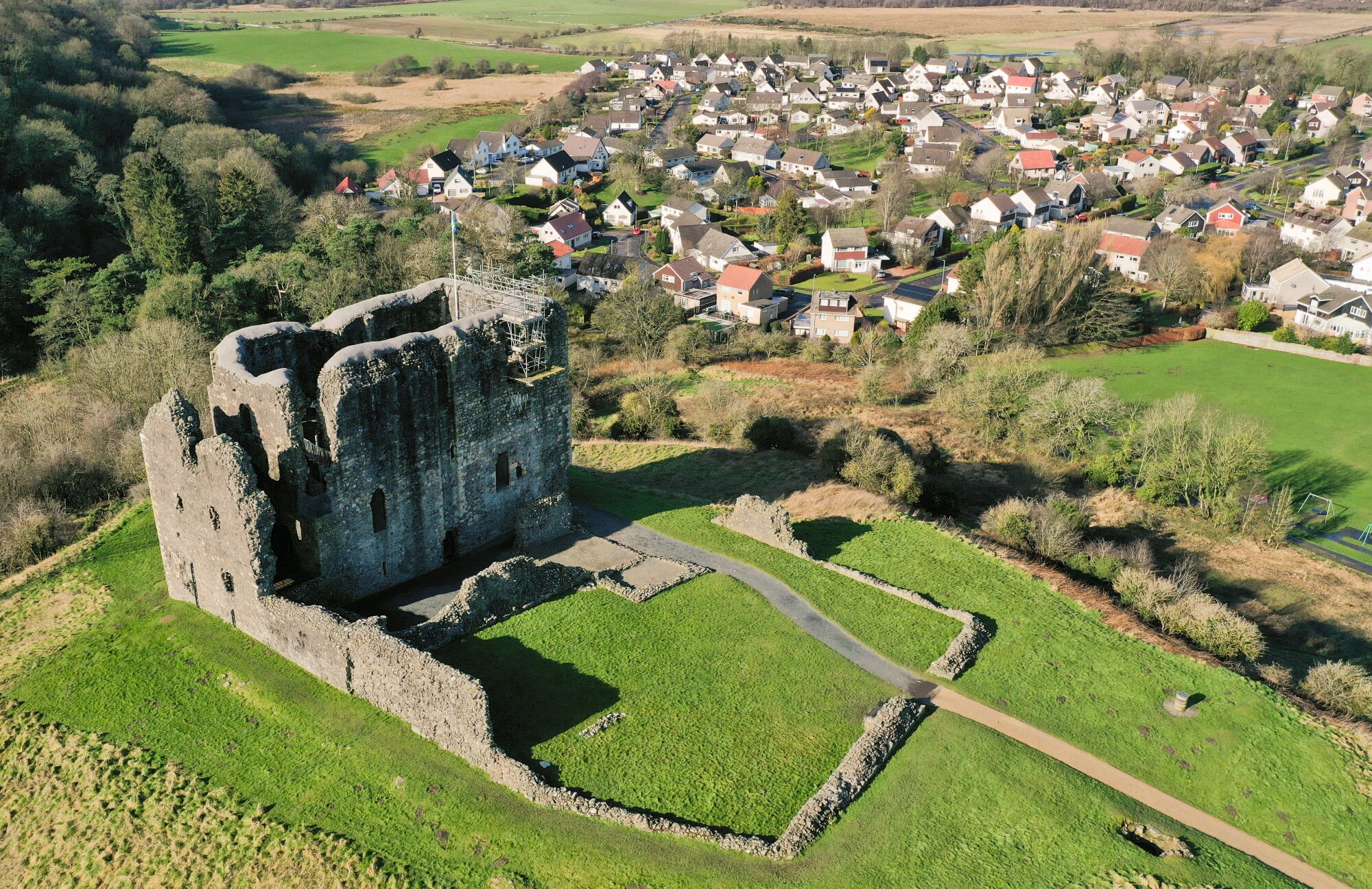 A ruined castle on a hill, with a modern housing estate in the background. The castle is a tall rectangular grey building without a roof surrounded by a low wall.