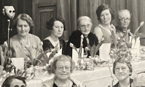 Groups of women at some kind of reception sit at tables decorated with flowers. There is a row of five women of varying ages in the rear of photo, along a straight table. 