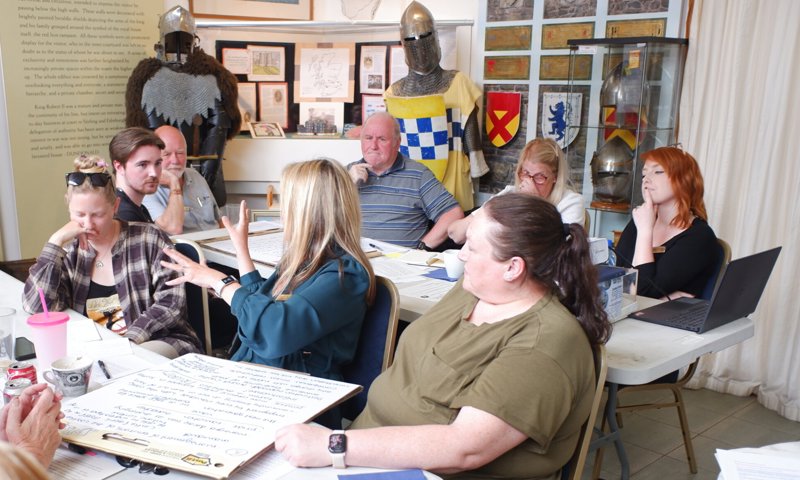 A group of people sitting around two tables in a room discuss things written on a flipboard lying flat on the table. One of them is talking with raised arms and the others are listening. In the background is a display of castle items and mannequins dressed as knights.
