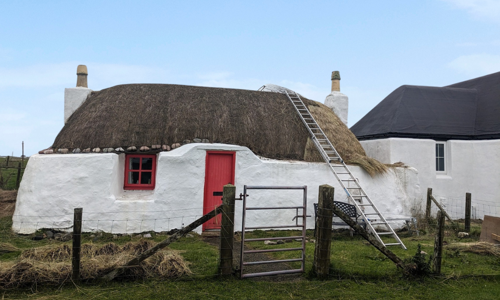 A small stone built, thatched, lime washed cottage with a red front door. A ladder leans on the roof and it looks like the cottage is half way through being re-thatched.