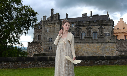 A lady wearing a long cream dress bolero jacket holding letters with Stirling Castle's Royal Palace behind her