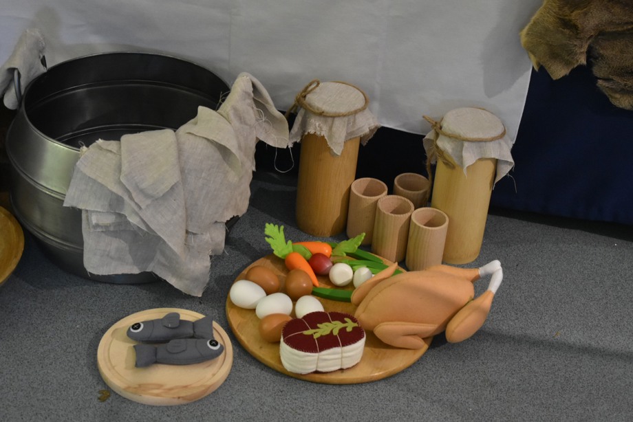 Items from the pack a cauldron kit: a metal cauldron; wooden cups and storage vessels; plastic children's food items including a roast chicken, fish, eggs, a cut of meat, carrots and other vegetables.