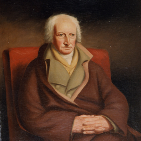 Image is a colour portrait painted of a person sitting in a chair with their hands in their lap, fingers interlaced. They are wearing several layers with a brown coat, and are looking with a serious expression to the right of the painter.