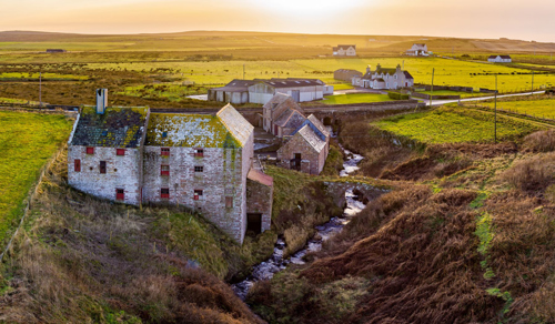 Aerial photo of John O'Groats mill and surrounding landscape. A small stream runs alongside various buildings, the largest of which has its roof tiles covered in yellow moss. It is sunset.