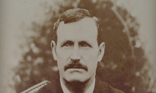 Sepia toned, front on portrait photo of gentleman in a suit, with a large moustache and short black hair parted on the side.