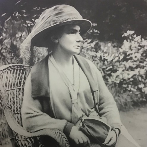 Black and white image of woman sat in wicker chair in a garden. She is wearing a hat and holding a handbag on her lap.