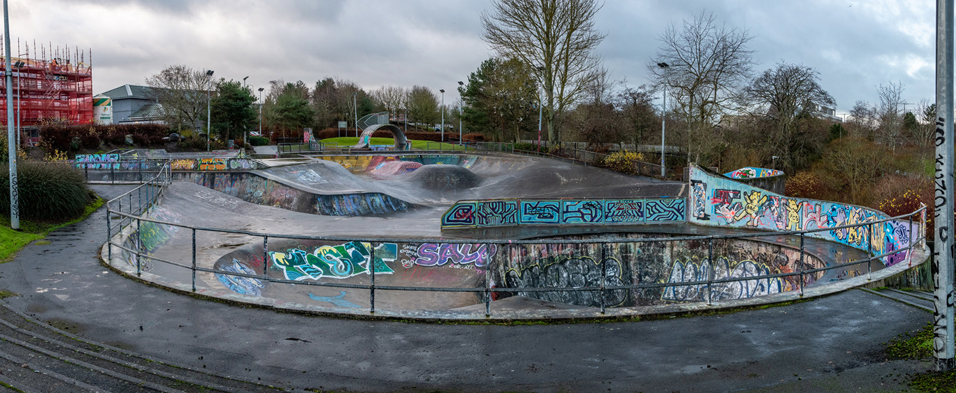 Livingston skatepark on a wet, winter day. The concrete is darkened from rain, and the tree around the park are devoid of leaves. The skatepark itself is covered in colourful graffiti, and nearest our view is two skatebowls shaped like the number eight.