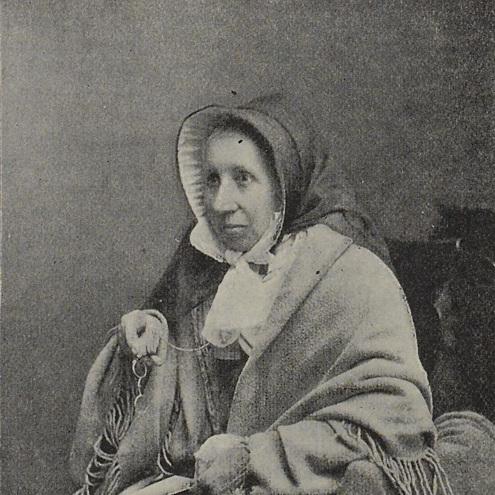 Sepia toned, older newspaper image of woman sat for portrait photo facing at a 45 degree angle. She is wearing period clothing, appears to be wrapped in a shawl and holds her round metal glasses in her right hand.